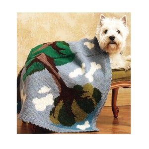 May include: A white dog with a blue, green, and brown knitted blanket with a tree and bone designs. The dog is lying on a light brown wooden bench.
