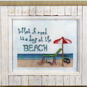 以下が含まれることがあります： 「What I need is a day at the BEACH」というテキストと、ビーチチェア、パラソル、海を描いたビーチシーンが描かれた、額装されたクロスステッチデザインです。
