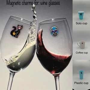 Może przedstawiać: Magnetyczne zawieszki do kieliszków do wina. Dwa kieliszki do wina z czerwonym i białym winem, każdy z ozdobną zawieszką. Jedna zawieszka to czarny kot, druga kolorowa czaszka. Dodatkowe zdjęcia pokazują kubek Solo, filiżankę do kawy i plastikowy kubek.