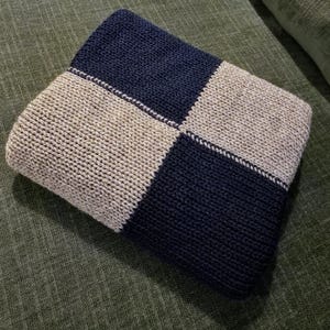 May include: Folded knitted blanket in a checkerboard pattern. The blanket features alternating squares of navy blue and beige. The knitted texture is visible, and the blanket appears soft and cosy. The blanket is folded neatly.