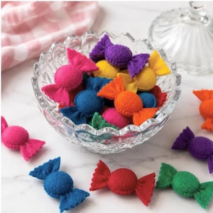May include: A collection of small, felted wool candies in various bright colors including pink, purple, yellow, orange, blue, red, and green. Several of these soft, wrapped-style felt candies are contained within a clear, scalloped glass bowl.
