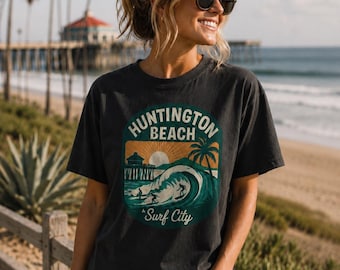 May include: A charcoal gray short-sleeve t-shirt featuring a graphic print with the text Huntington Beach Surf City. The design depicts a sunset, a pier, a palm tree, and surfers riding a large ocean wave in shades of green, orange, and white.