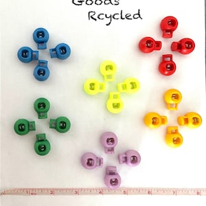 Puede incluir: Surtido de extremos de cordón de plástico de colores. Los topes están en seis colores: azul, verde, amarillo, naranja, morado y rojo. El texto "Random Goods Recycled" está escrito en la parte superior de la imagen.