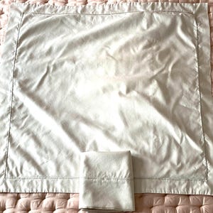 May include: A light beige pillowcase and a folded matching sheet set on a pink quilted surface. The pillowcase is rectangular with a stitched border. The sheet is also rectangular with a stitched border.