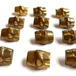 May include: A collection of 12 gold-toned metal buttons with a textured, raised design. The buttons are all the same size and shape, and they have a slightly rounded rectangular shape.
