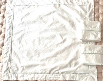 May include: A white bed sheet set with a subtle gray border. The set includes a large flat sheet and two pillowcases, all made of a soft, slightly wrinkled fabric. The sheets are neatly arranged on a textured, light pink surface.