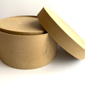 May include: A round, cardboard box with a lid slightly ajar. The box and lid are made of a light brown, uncoated material. The box is cylindrical, and the lid is a flat circle. The box is sitting on a white surface.