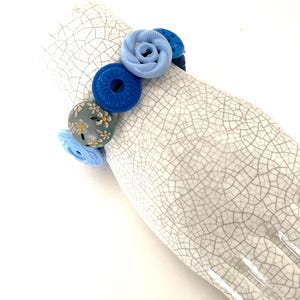 May include: A bracelet composed of various circular buttons in shades of blue, light blue, and a grey button featuring a white and yellow floral pattern. The buttons are strung together, displaying different textures, embossed designs, and center holes.