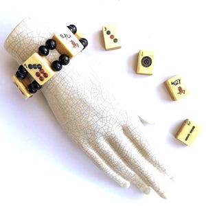 Puede incluir: Un brazalete vintage hecho de fichas de Mahjong de color marfil y cuentas negras. El brazalete se exhibe en una mano de cerámica blanca, con varias fichas sueltas cerca. Las fichas presentan marcas rojas y negras.
