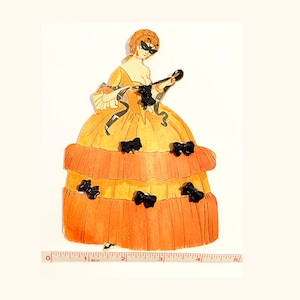 May include: A paper doll of a woman wearing a yellow and orange dress with black bows. The doll is holding a black mask in her right hand.