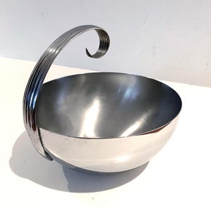 May include: A polished silver-toned metal bowl with a smooth, rounded interior and a single, tall, curved handle. The handle features a ribbed, textured surface and terminates in a decorative, tight spiral curl that extends above the bowl rim.