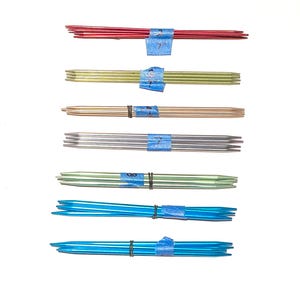 May include: A collection of knitting needles in various colors, including red, gold, silver, green, and blue. Each set is bundled with blue tape and a black rubber band. The needles are arranged against a white background.
