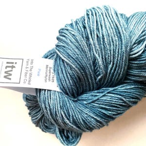 Peut inclure: Un écheveau de fil bleu avec une texture variée et une étiquette en papier blanc. L'étiquette porte le texte 'itw', 'Into The Whirled Yarn & Fiber Co.', et 'Pixie', ainsi que '75/25 Superwash Merino/Nylon' imprimé à l'encre noire sur papier blanc.