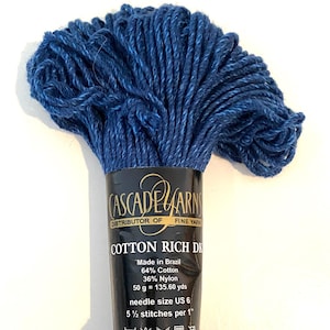 Op de afbeelding: Een streng diepblauw garen met een zwart label met de tekst Cascade Yarns, Cotton Rich DK, Made in Brazil, 64% Cotton, 36% Nylon, 50 g = 135,60 yds, needle size US 6, 5 1/2 stitches per 1 inch en vijf wassymbolen aan de onderkant.