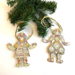 Op de afbeelding: Twee decoratieve kerstversieringen in de vorm van figuren. De ene heeft een kleurrijk ontwerp met een plank met items, de andere is versierd met klokgezichten. Beide hebben touwhangers en staan in de buurt van een groene dennentak.