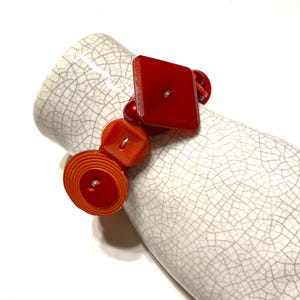 Puede incluir: Un brazalete de cuentas rojas y naranjas con botones grandes y coloridos. El brazalete está hecho con una variedad de formas y tamaños de botones, incluyendo un botón cuadrado, un botón redondo y un botón con un diseño en espiral.