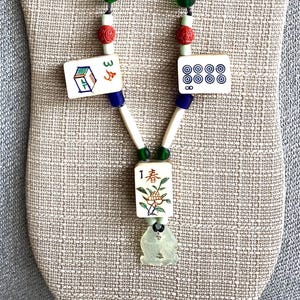 May include: A necklace featuring three off-white Mahjong tiles with colorful symbols and characters, including the number 1 and a floral design. The piece is accented with red, blue, green, and cream-colored beads and a pale green carved pendant.