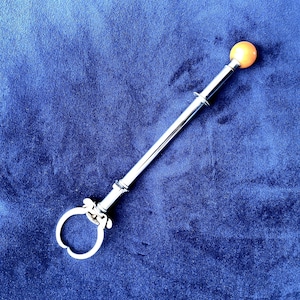 Puede incluir: Un utensilio de servir plateado con una bola de color amarillo anaranjado en un extremo y un asa circular en el otro. El utensilio se encuentra sobre un fondo de terciopelo azul oscuro.