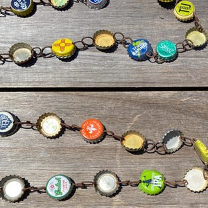 May include: A decorative garland constructed from various colorful metal bottle caps linked together with small dark brown metal rings. The caps feature diverse brand logos and designs in colors including yellow, blue, green, orange, and white.