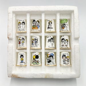 Könnte beinhalten: Ein Set aus zwölf Keramik-Fingerhüten mit goldenem Rand, die jeweils eine andere Illustration des Comics "Love is..." zeigen. Die Fingerhüte sind in einer weißen Schaumstoffschale angeordnet.