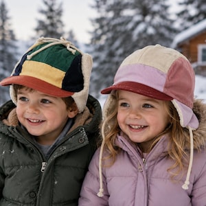 May include: Two corduroy earflap hats. The left hat features a color-blocked design in green, yellow, navy, and rust with cream-colored lining. The right hat features a color-blocked design in cream, light pink, and mauve with a dark red brim.