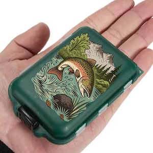 Może przedstawiać: Ciemnozielone pudełko na sprzęt wędkarski z szczegółową ilustracją pstrąga wyskakującego z wody, otoczonego sceną gór i lasu. Pudełko ma czarne zapięcie i kilka małych przegródek.