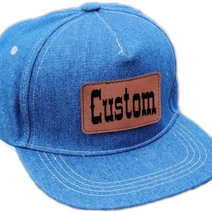 Peut inclure: Une casquette de baseball en denim bleu avec un patch en cuir marron qui dit "Custom" en lettres noires.