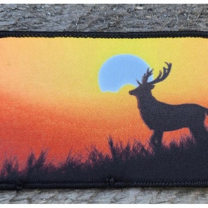 Könnte beinhalten: Eine schwarze Silhouette eines Hirsches mit Geweih, das in einem Feld bei Sonnenuntergang steht. Die Sonne ist leuchtend gelb und orange mit einem blauen Zentrum.