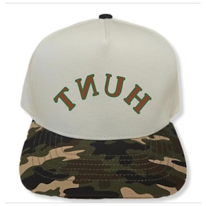 May include: A white baseball cap with a camouflage brim. The front of the cap has the word "TIUH" written in brown letters with a green outline.