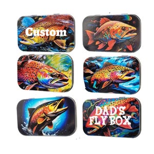 May include: Six colorful metal fly boxes with different designs. Each box features a trout with vibrant colors and a unique design. The boxes include designs with the words "Custom" and "Dad's Fly Box".