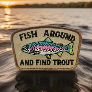 Puede incluir: Un parche rectangular con el texto "FISH AROUND AND FIND TROUT" encima y debajo de una ilustración de trucha colorida. La trucha presenta detalles azules, verdes, rosas y amarillos. El parche tiene un fondo beige y un borde marrón.