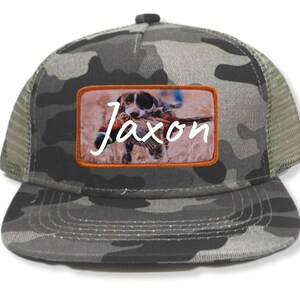 May include: A camouflage trucker hat with a brown patch featuring a dog with a pheasant in its mouth. The patch has the name "Jaxon" written in white cursive.