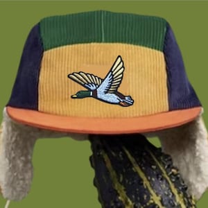 Puede incluir: Un gorro de pana multicolor con orejeras. El gorro presenta un panel superior verde, un panel central amarillo con un parche de pato volador, paneles laterales azul marino y una visera naranja. Las orejeras están forradas con un material afelpado de color crema.