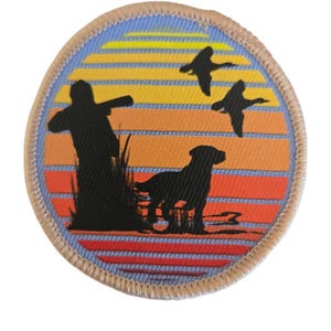 Op de afbeelding: Ronde patch met een jachtscène met een jager, hond en vliegende eenden tegen een zonsondergang. De patch heeft een bruine rand en een blauw, oranje en geel kleurenschema.