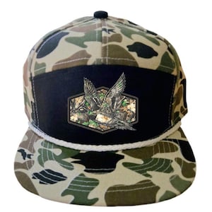 Könnte beinhalten: Camouflage-Baseballkappe mit schwarzem Frontpanel und weißem Seildetail. Die Kappe hat einen Aufnäher mit einer detaillierten Illustration von zwei fliegenden Enten vor einem Camouflage-Hintergrund. Das Muster der Kappe umfasst Grün-, Braun- und Beigetöne.