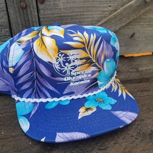 Könnte beinhalten: Eine blaue und weiße Baseballkappe mit tropischem Blumenmuster. Die Kappe hat einen weißen Kordelbesatz und ein Logo für "Special Olympics Kansas" auf der Vorderseite.