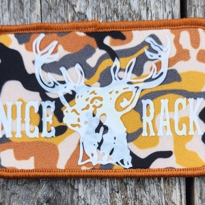 Könnte beinhalten: Ein brauner, schwarzer und weißer Tarnstoff-Patch mit einer weißen Hirschkopf-Silhouette und dem Text "NICE RACK" in Weiß.
