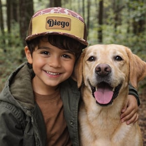 Op de afbeelding: Een bruin en beige truckerpet met de naam "Diego" op een leren patch. De pet heeft een mesh achterkant en een camouflagepatroon aan de voorkant. De pet wordt gedragen door een lachend kind. Een gele Labrador Retriever-hond staat naast het kind.