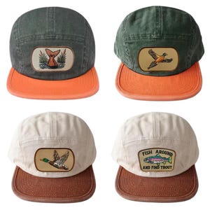 Może przedstawiać: Cztery płócienne czapki z różnymi wzorami. Dwie czapki są w kolorze oliwkowym z pomarańczowymi daszkami, z naszywkami z rybim ogonem i bażantem. Pozostałe dwie są w kolorze złamanej bieli z brązowymi daszkami, jedna z kaczką, a druga z pstrągiem.