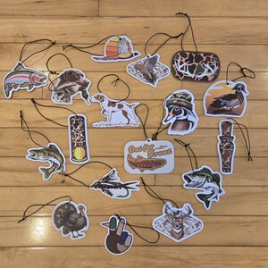 May include: A collection of air fresheners with hunting and fishing themes. The designs include fish, birds, dogs, and a deer. One air freshener has the text "Out for Trout". Each air freshener has a black string for hanging.