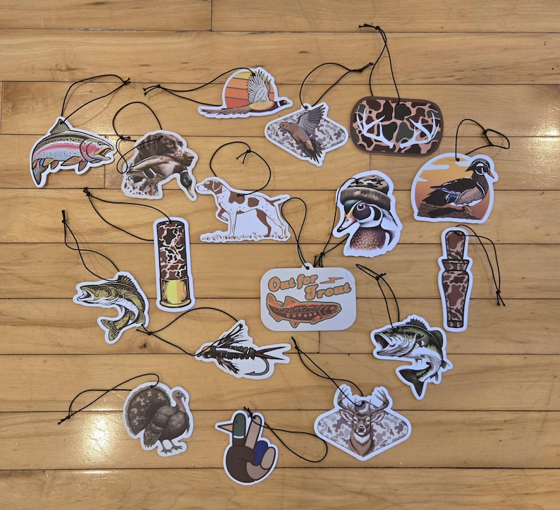 May include: A collection of air fresheners with hunting and fishing themes. The designs include fish, birds, dogs, and a deer. One air freshener has the text "Out for Trout". Each air freshener has a black string for hanging.