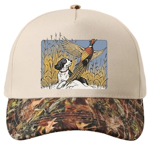 Puede incluir: Gorra de béisbol beige con visera de camuflaje. La gorra presenta un gráfico rectangular de un perro y un faisán en un campo. El perro es blanco y negro, y el faisán tiene plumas marrones, rojas y amarillas.