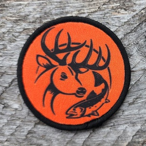 Könnte beinhalten: Orangefarbener und schwarzer gestickter Aufnäher mit einem Hirschkopf und Geweih sowie einem Fisch im Maul.