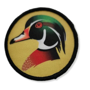 Puede incluir: Un parche redondo con borde negro que muestra una imagen colorida de la cabeza de un pato de madera. El pato tiene la cabeza verde, los ojos rojos, una franja blanca y el cuerpo rojo y negro.