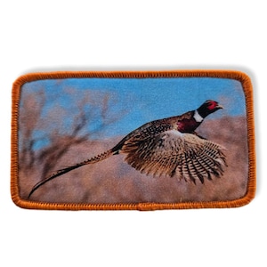 Könnte beinhalten: Ein braunes und weißes Rebhuhn mit rotem Kopf und Hals fliegt mit ausgebreiteten Flügeln in der Luft. Das Rebhuhn befindet sich vor einem blauen Himmel mit braunem Gestrüpp im Vordergrund. Das Bild ist auf einem braunen Stoffstück mit orangefarbenen Nähten an den Rändern.