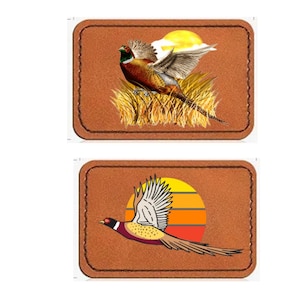 Puede incluir: Dos parches de cuero marrón con diseños de faisanes. El parche superior presenta un faisán volando en un campo de hierba alta con una puesta de sol en el fondo. El parche inferior presenta un faisán volando frente a una puesta de sol con rayas naranjas y rojas.