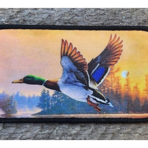 May include: A rectangular patch featuring a flying duck over a lake at sunset. The duck has green, blue, brown, and white feathers. The background shows a lake, trees, and a setting sun. The patch has a black border.