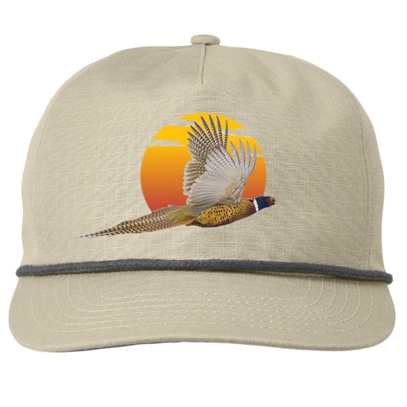 Pu&ograve; includere: Cappellino da baseball beige con una grafica di un fagiano in volo contro un tramonto. Il fagiano ha piume marroni, gialle e bianche. Il cappellino ha un dettaglio di corda grigia e una visiera curva.