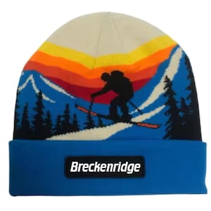May include: Blue knit beanie with a white and blue mountain scene and a skier silhouette. The beanie has a blue brim with a black patch that says "Breckenridge".