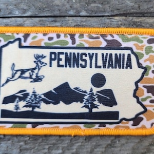Könnte beinhalten: Ein brauner, grüner und weißer Tarnstoff-Patch mit orangefarbenem Rand. Der Patch zeigt eine weiße Umrandung des Bundesstaates Pennsylvania mit einem Reh, das über eine Bergkette springt, und einer Sonne im Hintergrund. Der Text "PENNSYLVANIA" ist in schwarzen Buchstaben gedruckt.